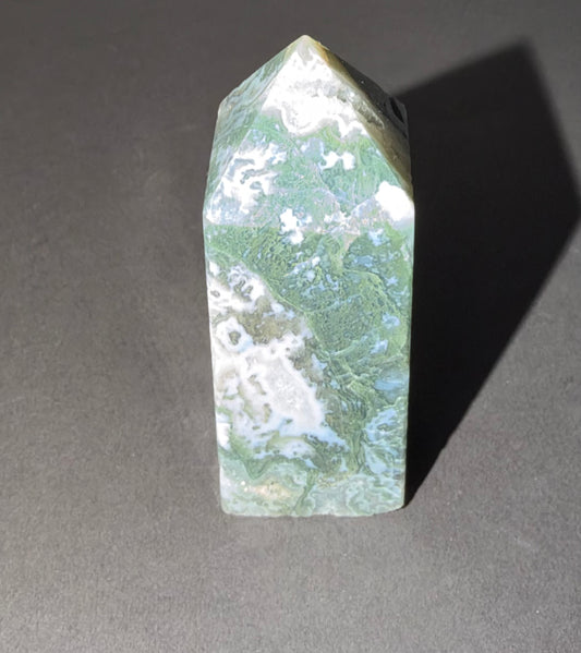 A polished natural crystal tower with green, white, and light blue marbling, standing upright on a dark gray surface with a sharp shadow.
