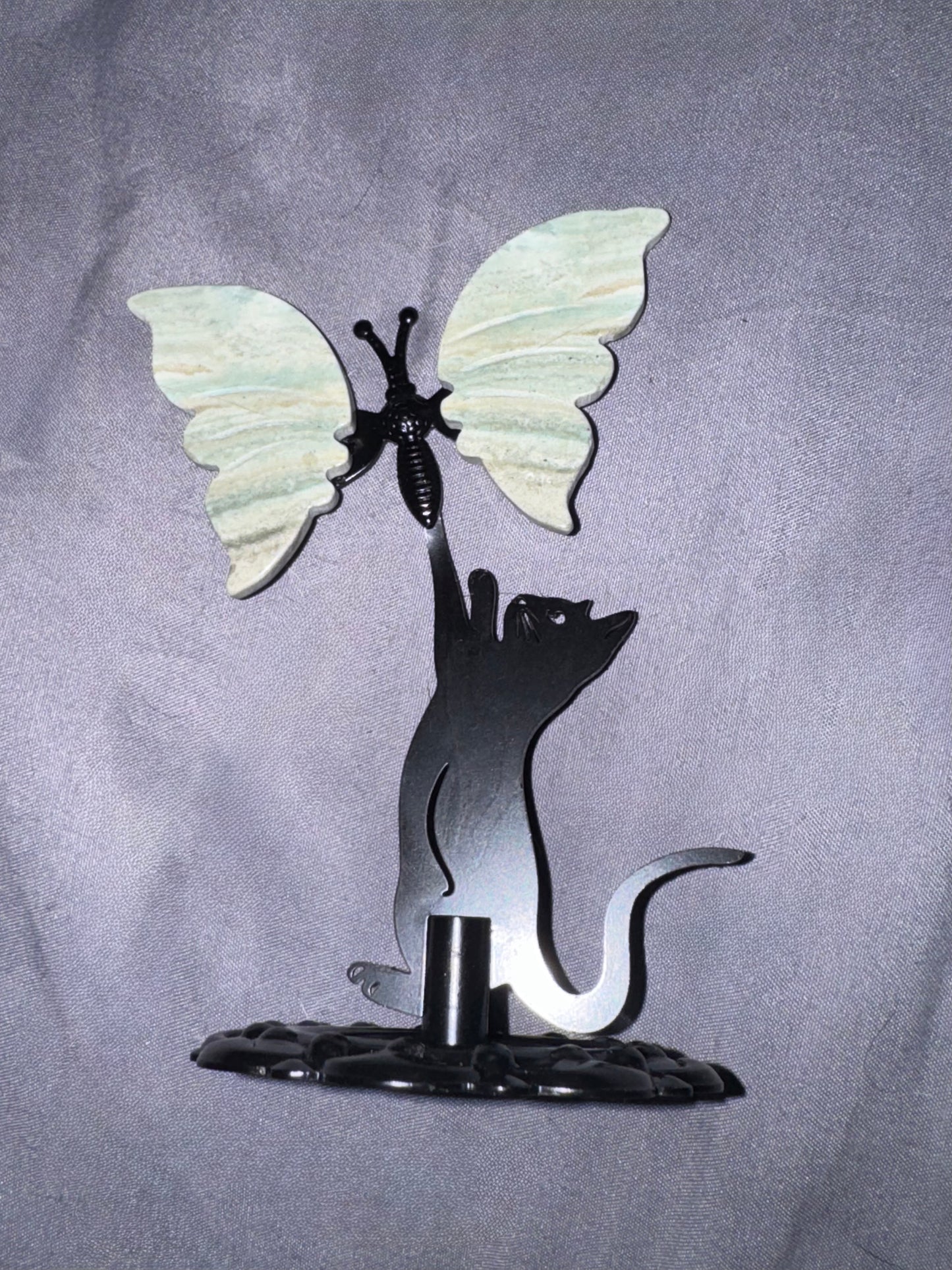 A decorative metal sculpture featuring a black cat silhouette reaching upward toward a butterfly with pale green and white wings, mounted on a dark base against a gray fabric backdrop.
