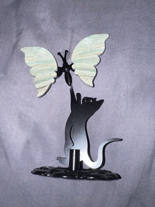 A decorative metal sculpture featuring a black cat silhouette reaching upward toward a butterfly with pale green and white wings, mounted on a dark base against a gray fabric backdrop.