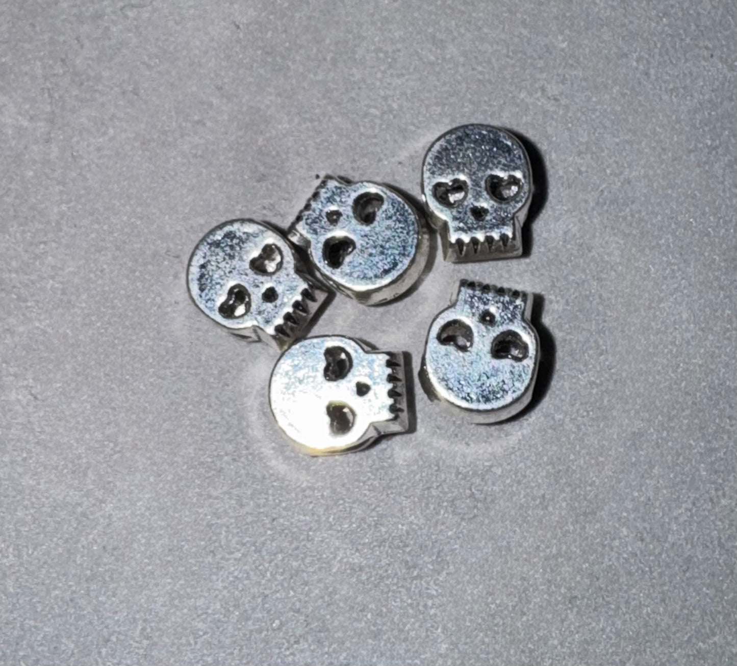 Five small silver skull-shaped beads or charms arranged on a textured gray surface.