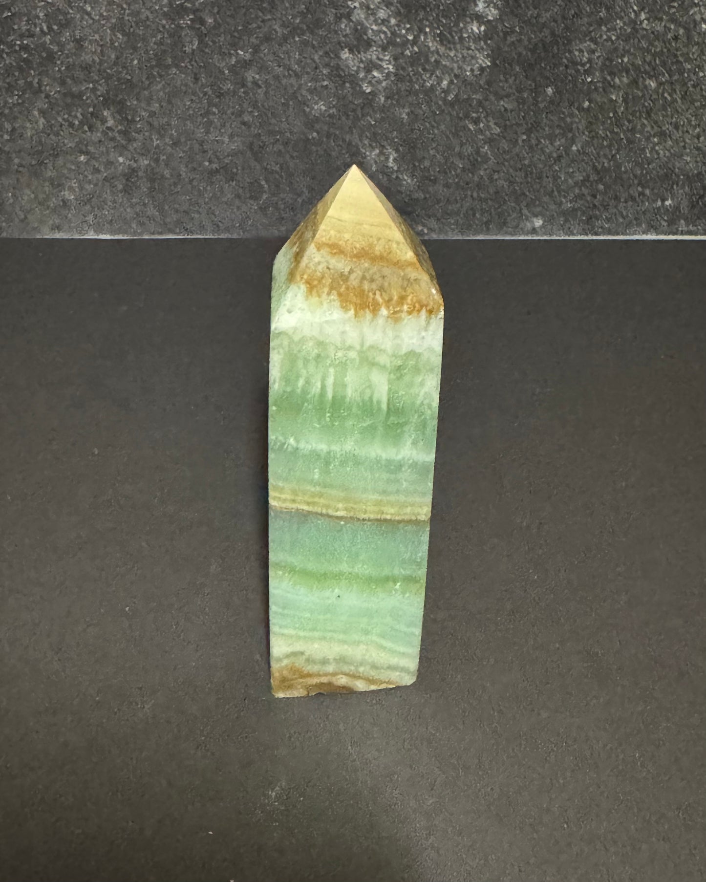 A polished mineral crystal tower with layered bands of light green, beige, and brown, standing upright on a dark gray surface.