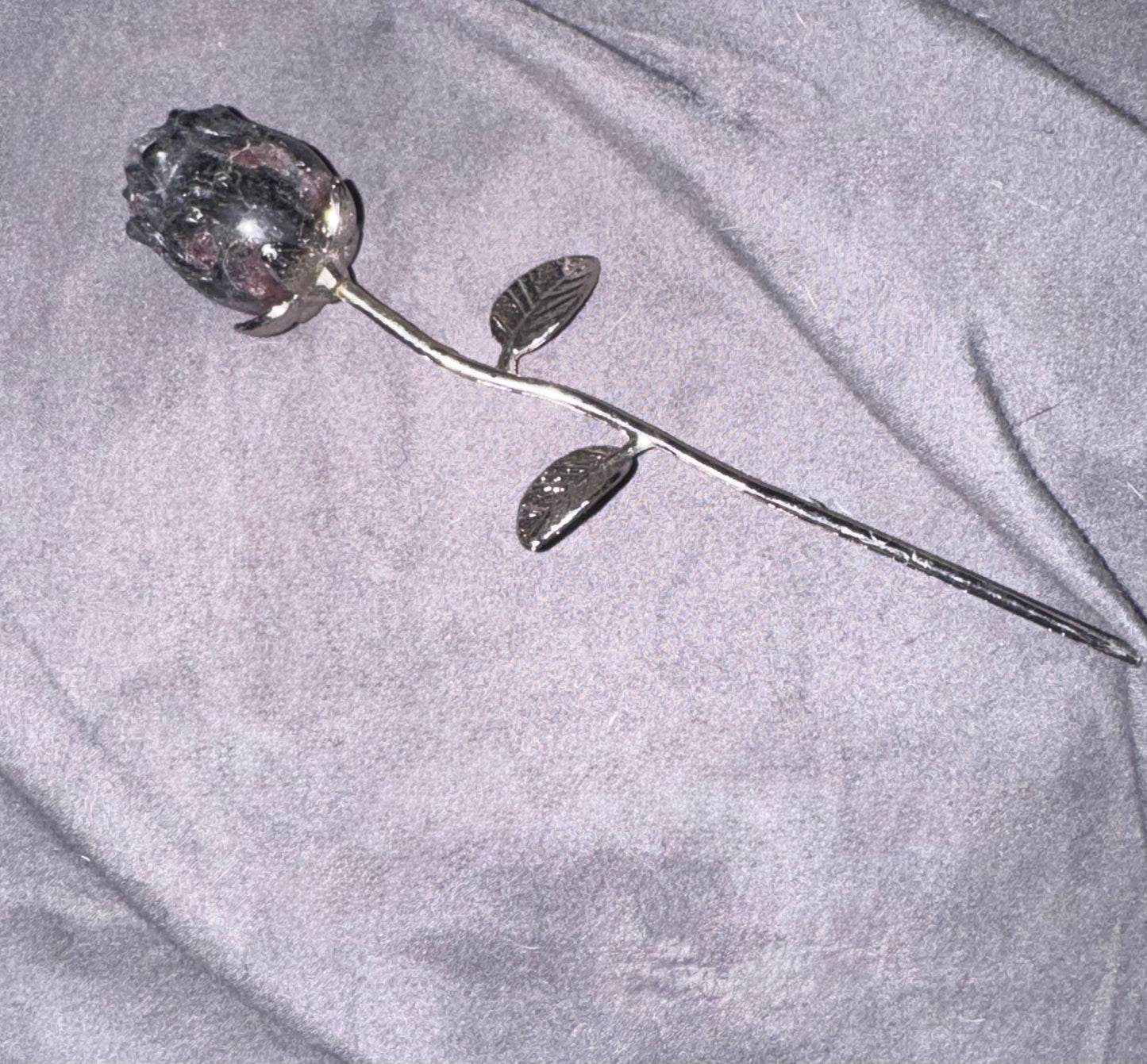 A single silver-toned metal rose stem with a faceted clear crystal rosebud and two leaves, placed on a wrinkled light gray fabric background.