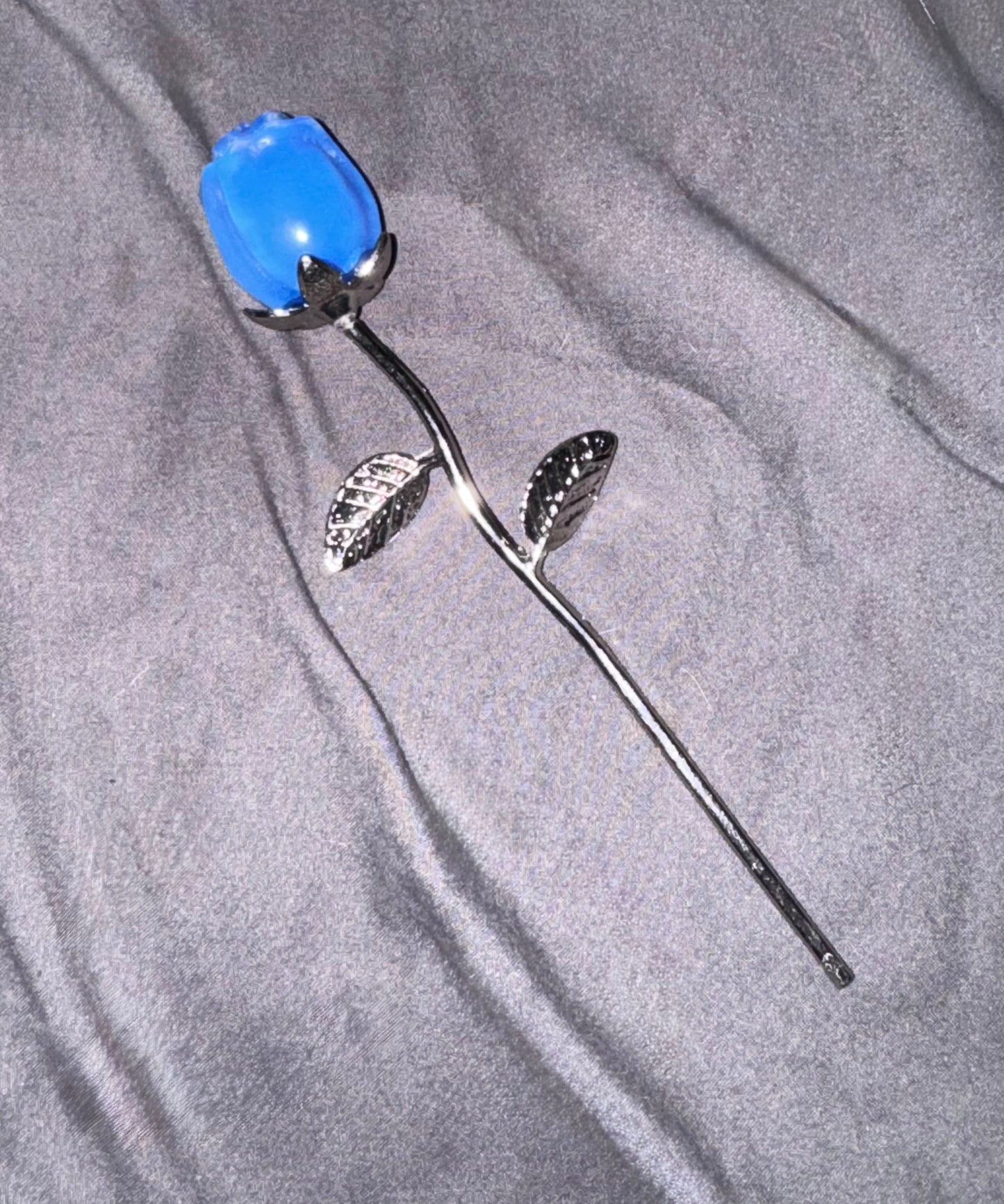A single artificial blue rose with a silver metal stem and textured leaves, placed on a gray fabric background.