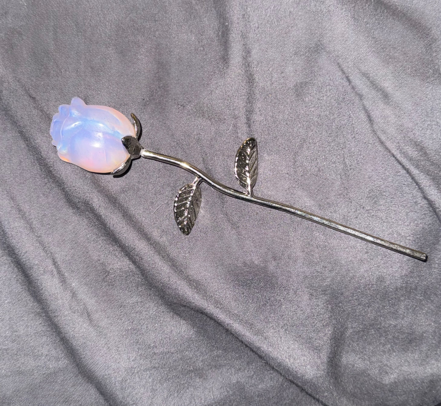 A silver metal rose- iridescent pale blue and pink opalescent rosebud, displayed on a textured gray fabric background.