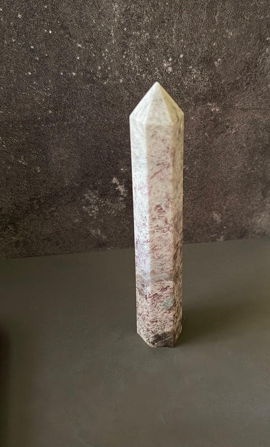A natural pointed crystal tower, standing upright on a dark gray surface against a textured dark gray wall.