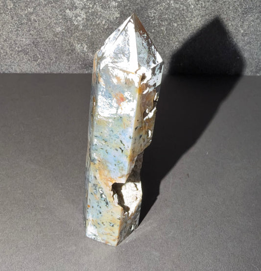 A natural silver-toned crystal point with iridescent blue, green, and orange mineral inclusions, standing on a dark gray surface against a textured gray background.