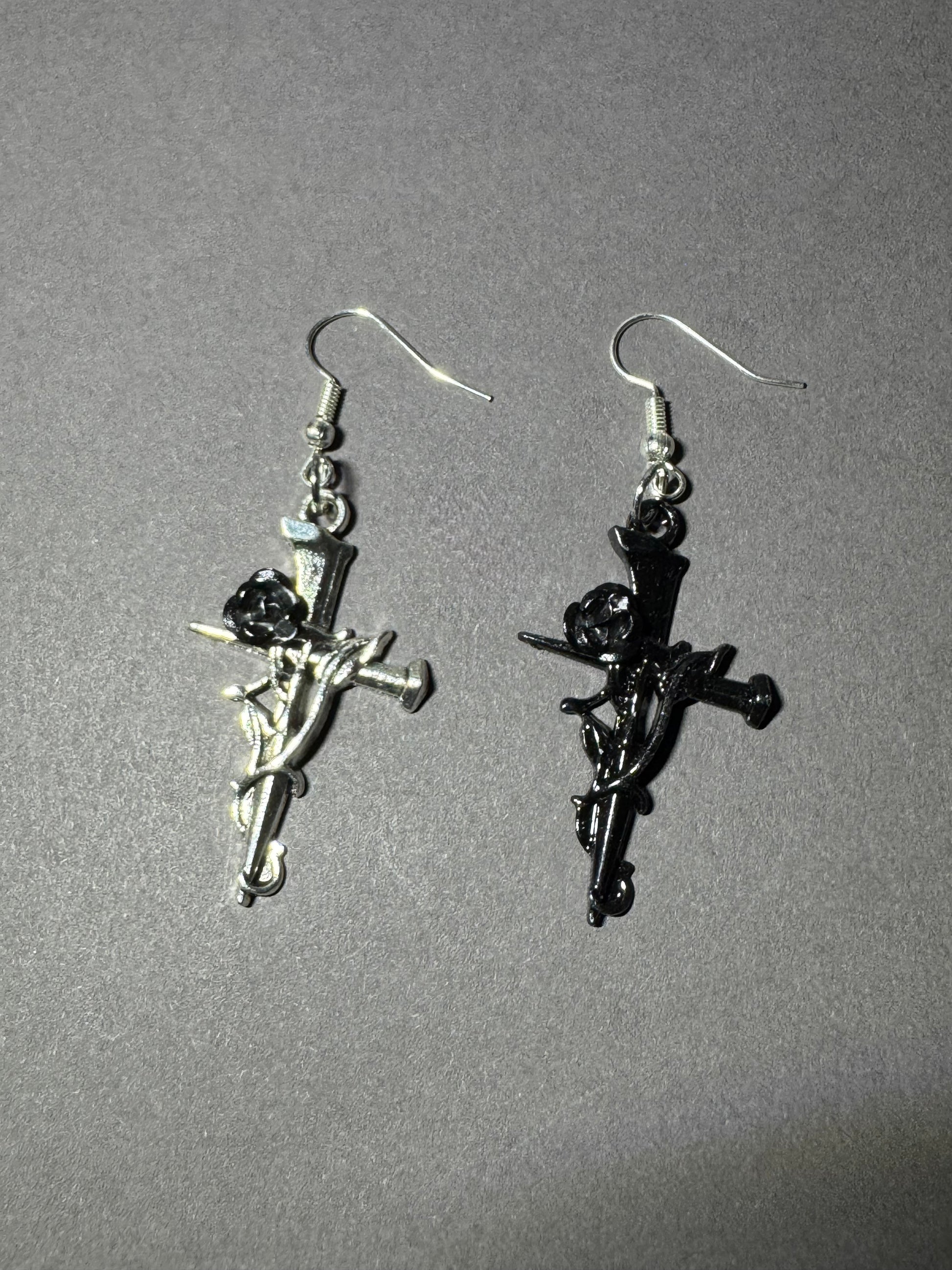 A pair of gothic-style dangle earrings featuring cherub figures on a sword, one in polished silver-tone metal and the other in black enamel, displayed on a neutral gray background.