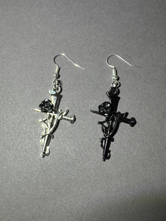 A pair of gothic-style dangle earrings featuring cherub figures on a sword, one in polished silver-tone metal and the other in black enamel, displayed on a neutral gray background.