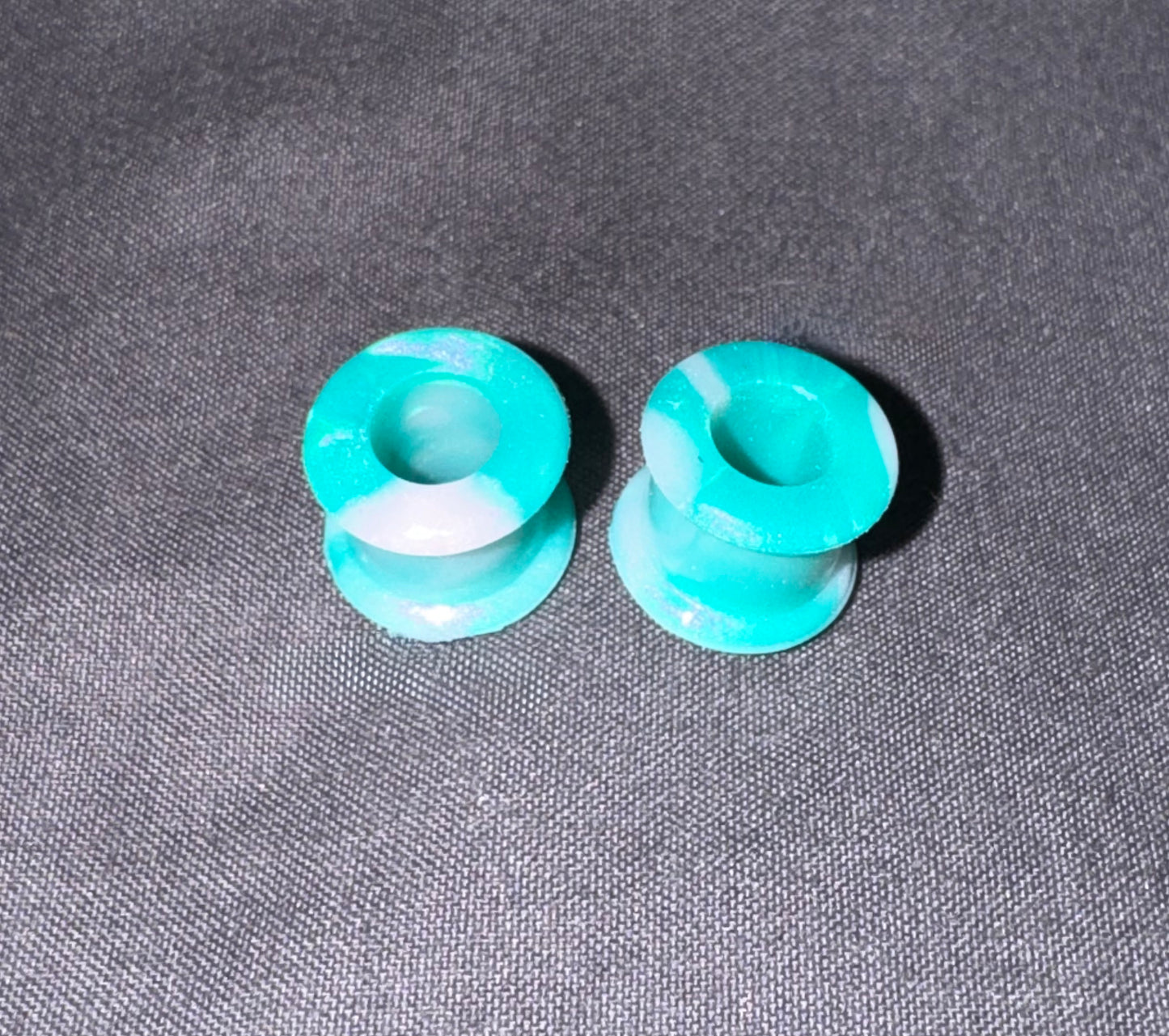 Two small turquoise blue and white marbled silicone plugs, likely for ear stretching or jewelry making, placed on a textured gray fabric background.