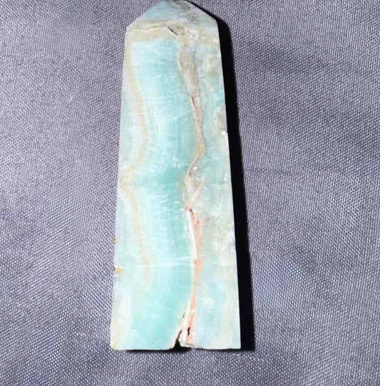 A polished natural stone or mineral point with a light aqua and white marbled pattern and beige veining, photographed on a textured gray fabric background.