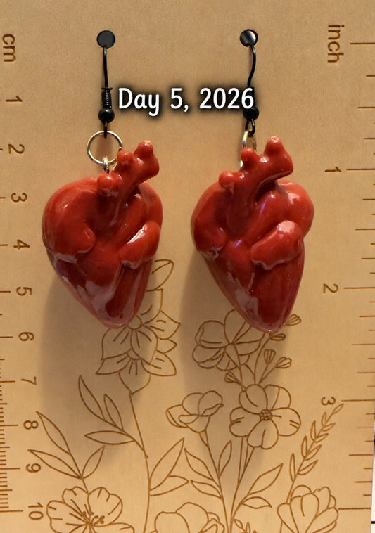 A pair of glossy red anatomical heart drop earrings displayed on a beige floral ruler background with measurement markings and the text 'Day 5, 2026' overlaid near the top.