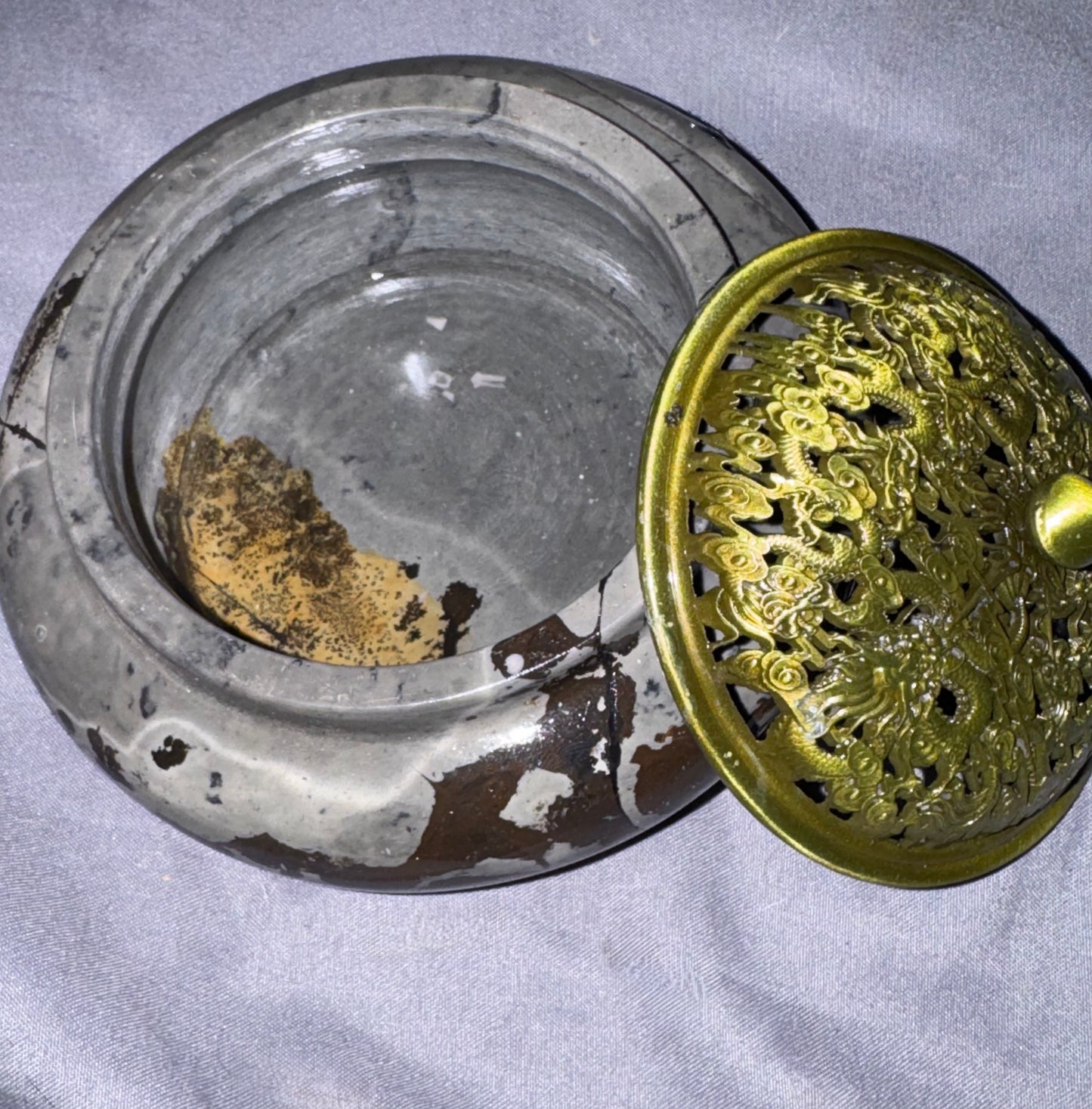 A round glass incense burner with a matching perforated metal lid featuring an ornate dragon design, resting on a light gray fabric background.