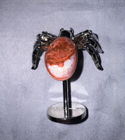 A spider with a polished red and white marbled cabochon stone on a  metal stand, set against a textured gray fabric background.