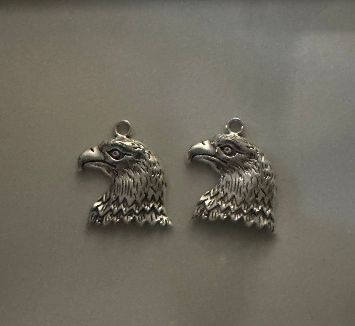 Two silver eagle head charms with detailed feather and beak textures, each featuring a small loop at the top, displayed on a neutral gray background.