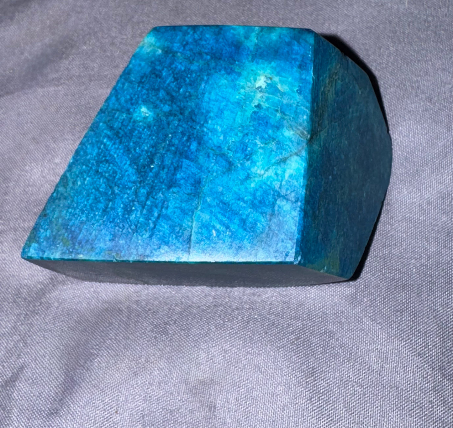 A faceted blue stone or crystal block with a polished finish, resting on a textured gray fabric surface.