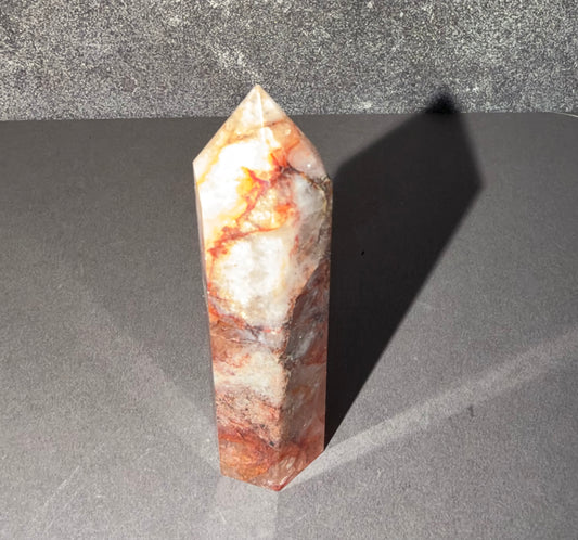 A natural quartz crystal point with orange, red, and white marbled patterns, standing upright on a gray surface with a sharp shadow cast to the right.