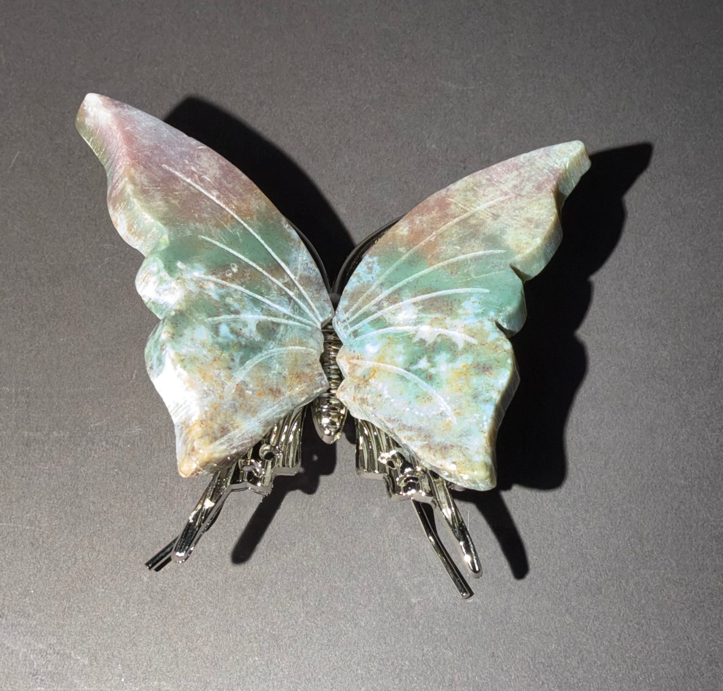 A butterfly-shaped ocean Jasper carving  featuring iridescent green, blue, and pink wings with silver metal legs, displayed on a dark gray background.