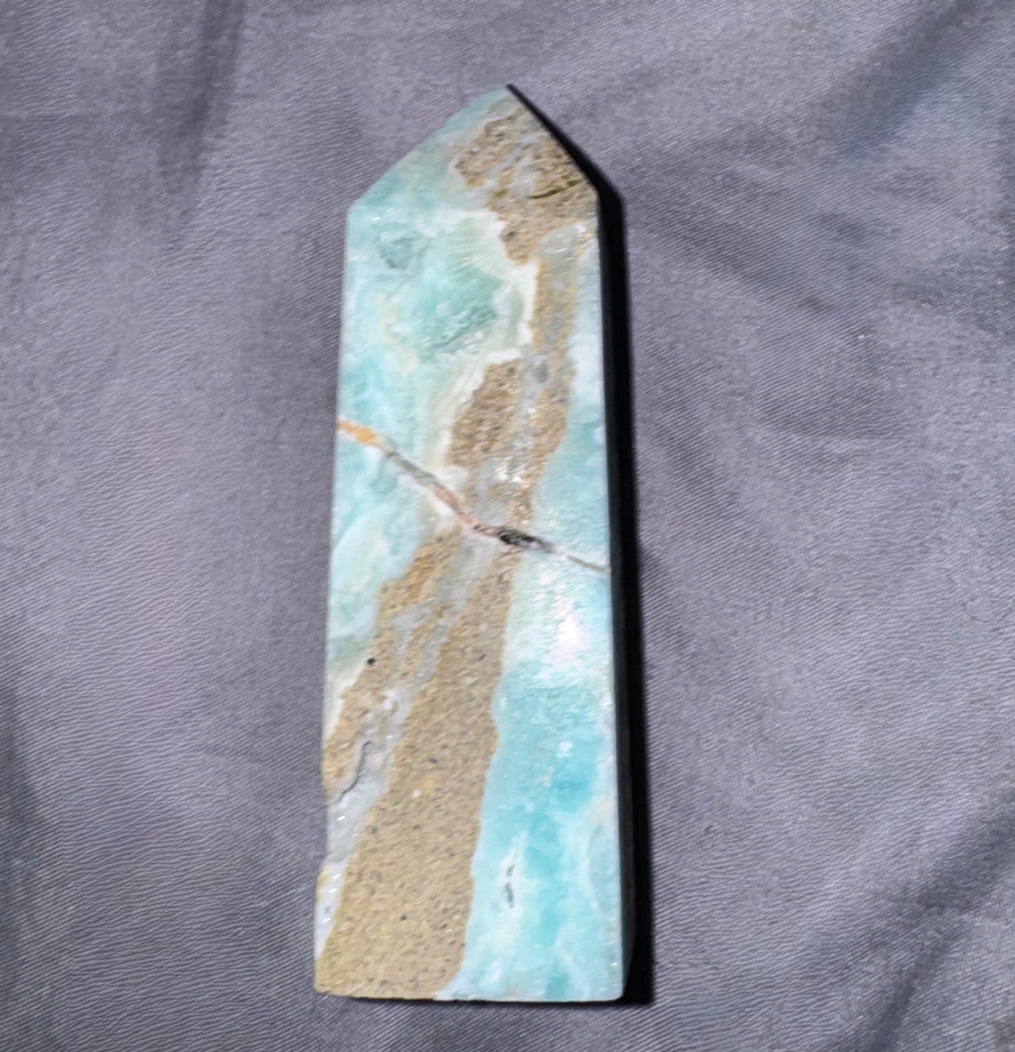 A polished pointed crystal tower made of light blue and white marbled stone with a prominent tan band, photographed standing upright on a gray fabric surface.