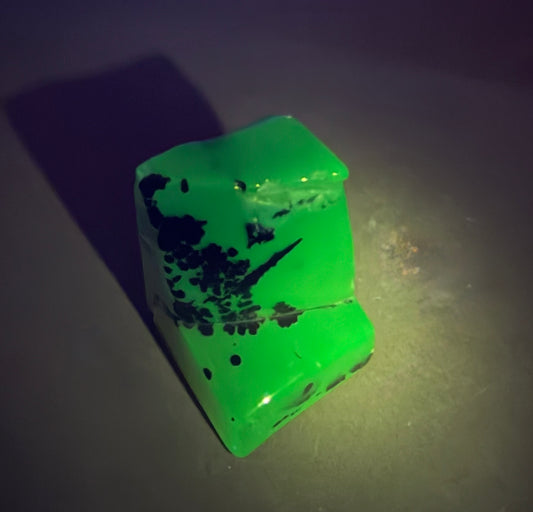 A translucent bright green cube-shaped gemstone or crystal with black inclusions and speckles, photographed on a dark surface with dramatic lighting.
