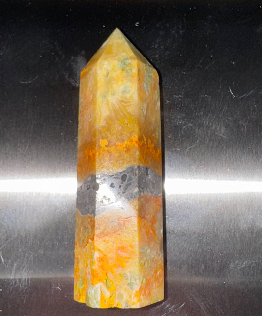 A natural crystal tower with a pointed top, featuring marbled patterns of orange, yellow, and a dark gray band in the middle, set against a reflective metallic background.