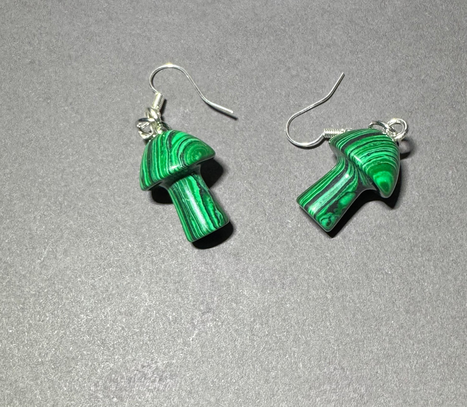 A pair of mushroom-shaped drop earrings made from green malachite with silver fishhook wires, displayed on a textured gray background.