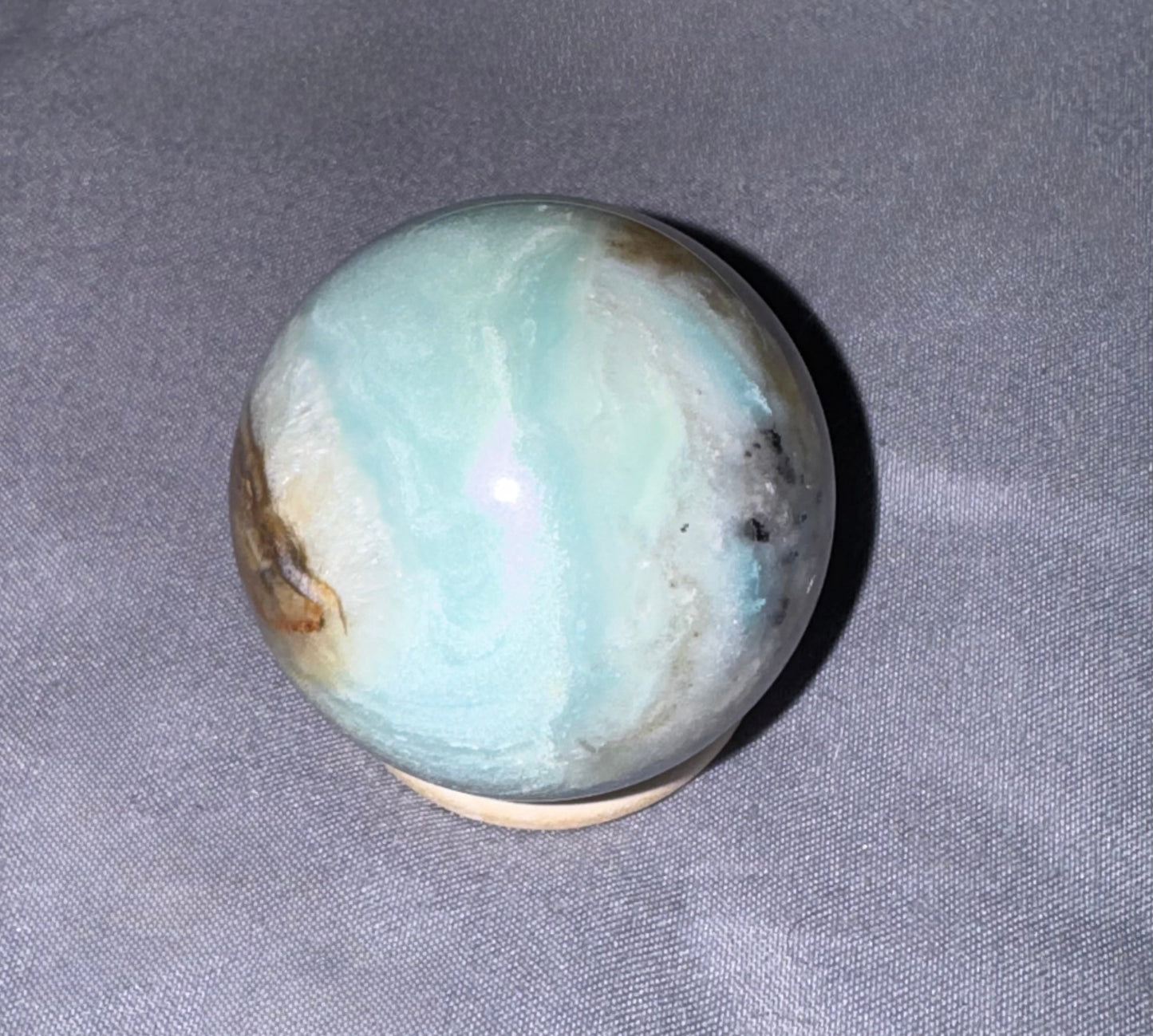 A polished Amazonite crystal sphere with light blue-green and white marbling, resting on a small wooden stand against a gray fabric background.