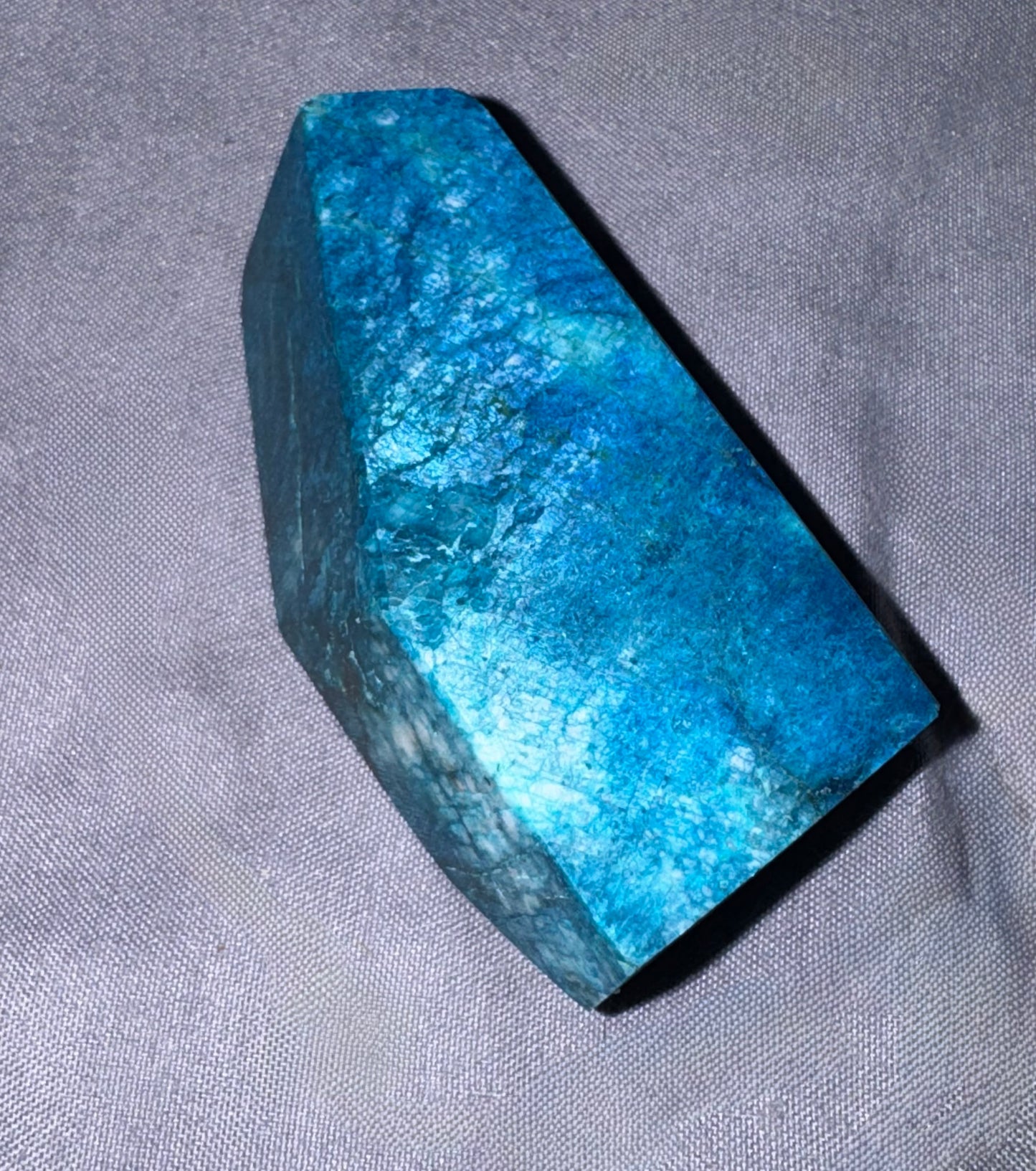 A polished blue stone or crystal with a faceted, geometric shape, resting on a textured gray fabric background.