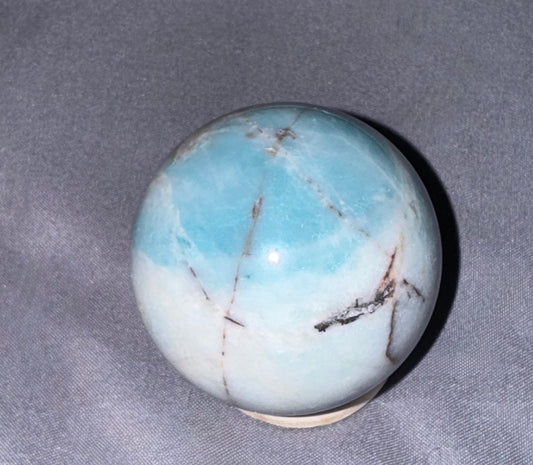 A polished light blue and white  crystal sphere with natural brown veining, displayed on a small beige base against a textured gray fabric background.