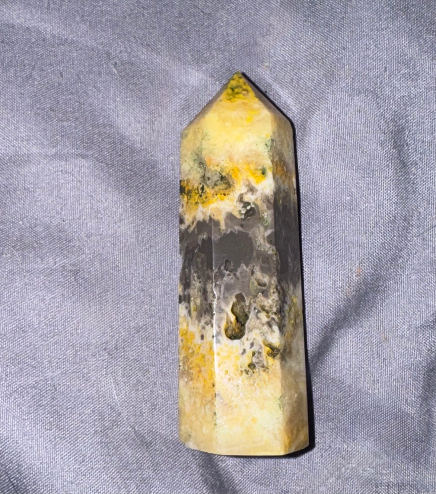 A faceted crystal tower or obelisk with a natural marbled pattern of yellow, black, and gray, resting on a textured gray fabric background.