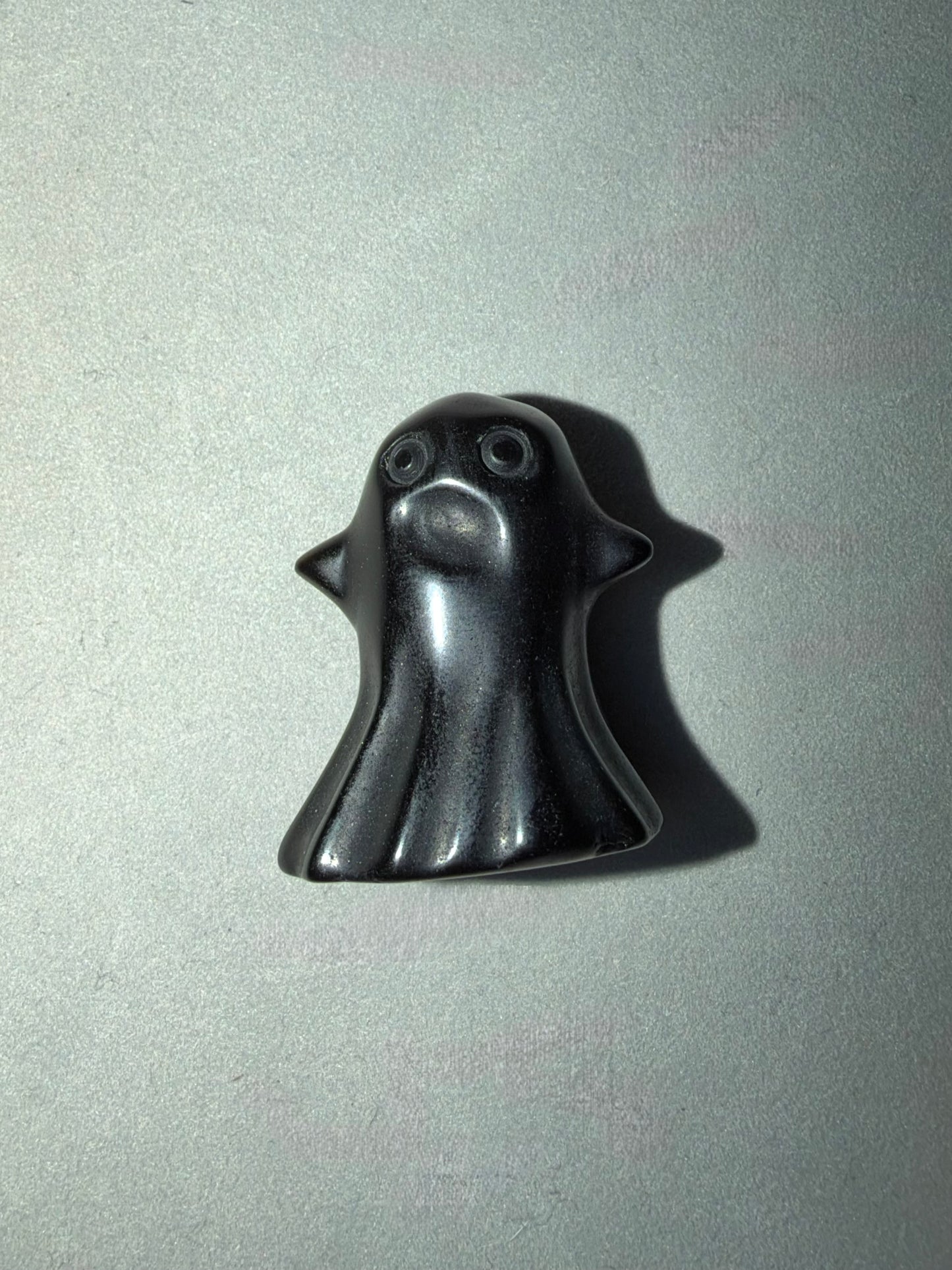 A small glossy black ghost-shaped object with simple facial features, photographed on a plain light gray surface with a cast shadow.