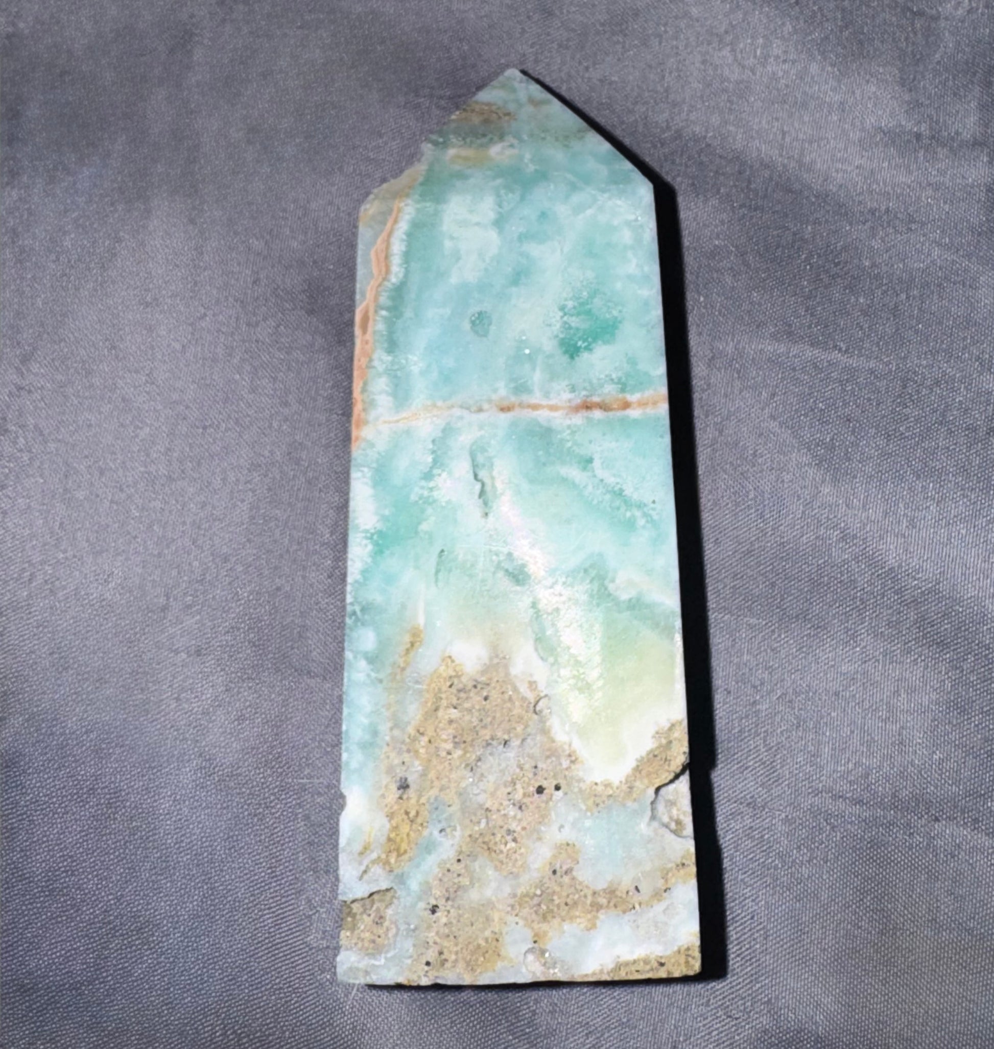 A natural pointed crystal tower or obelisk made of marbled aqua, mint green, and beige stone, photographed on a textured gray fabric background.
