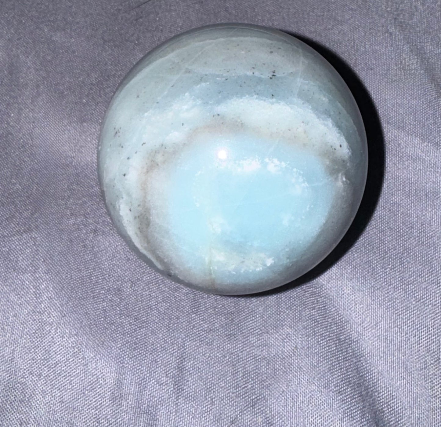 A polished light blue and white stone sphere, resembling a  crystal, resting on a textured gray fabric background.