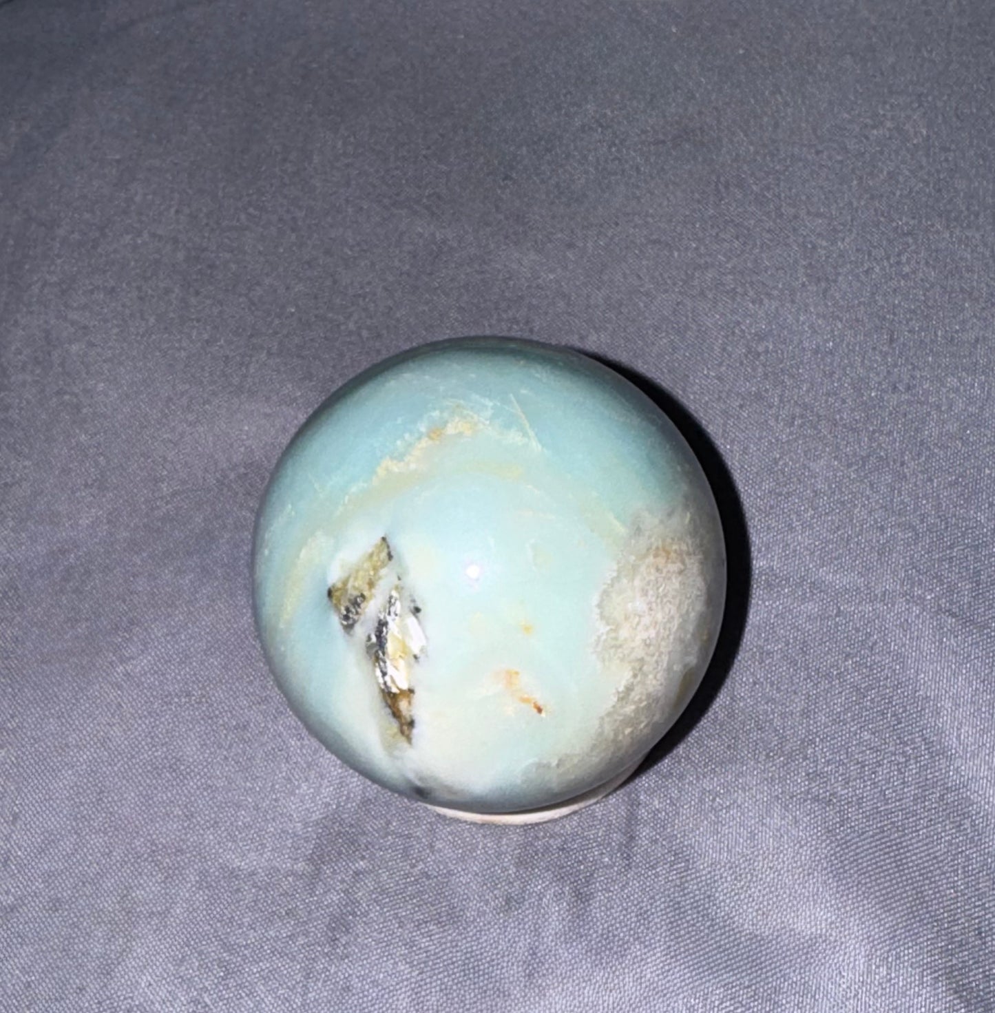A polished round stone sphere with a pale blue-green body, natural banding, and a metallic gold vein, resting on a gray textured fabric background.