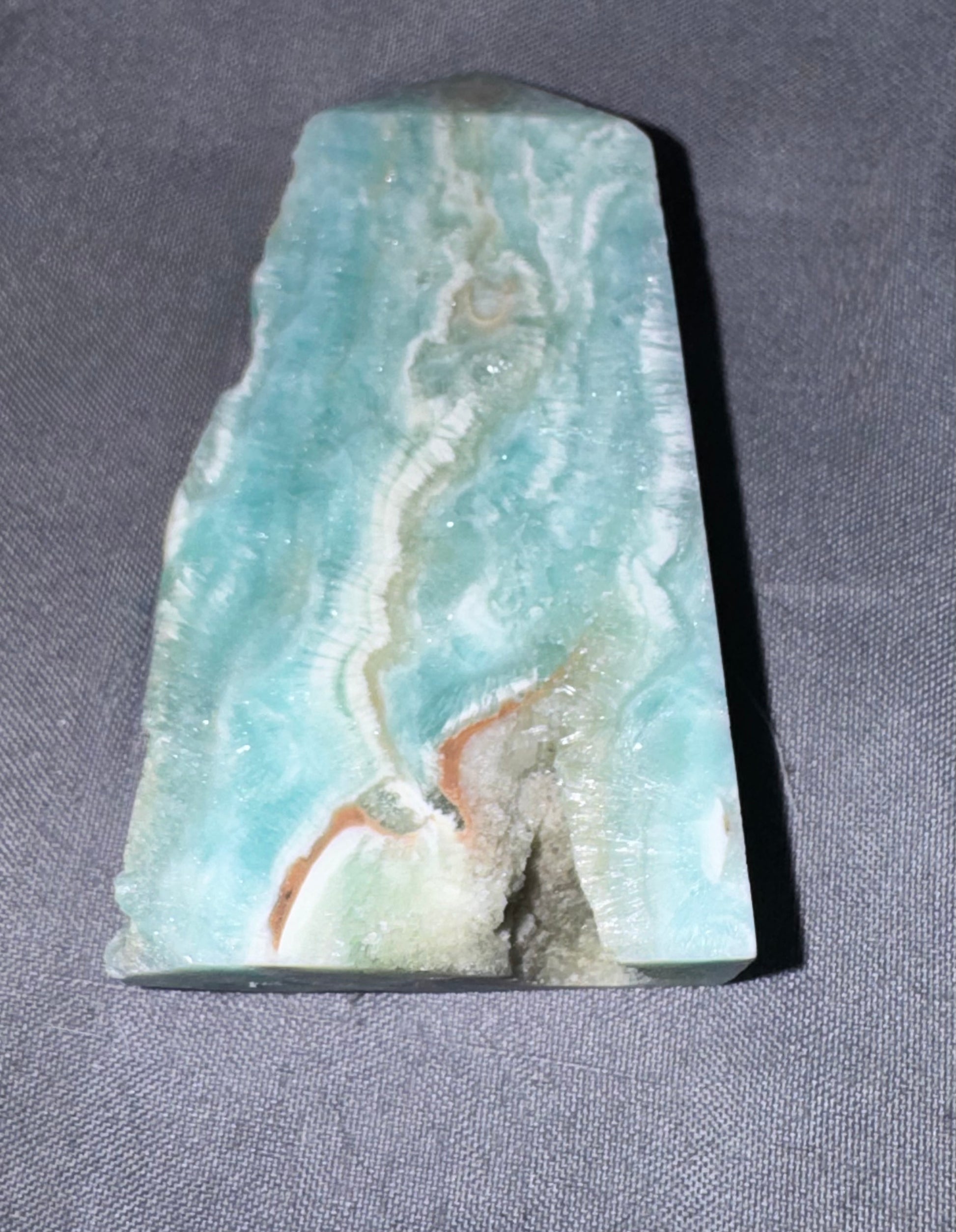 A polished natural stone slab with a marbled pattern of light blue, white, and soft orange-brown veins, resting on a textured gray fabric background.