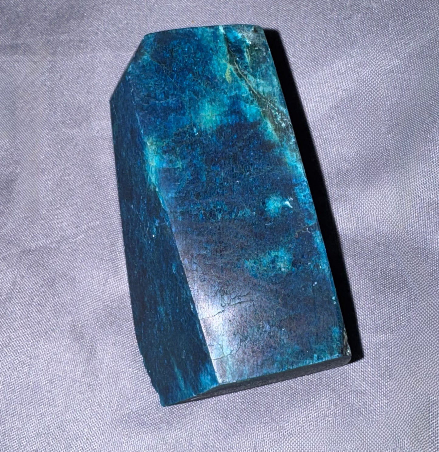 A faceted blue-green stone or crystal block with a polished, glossy surface, resting on a textured gray fabric background.
