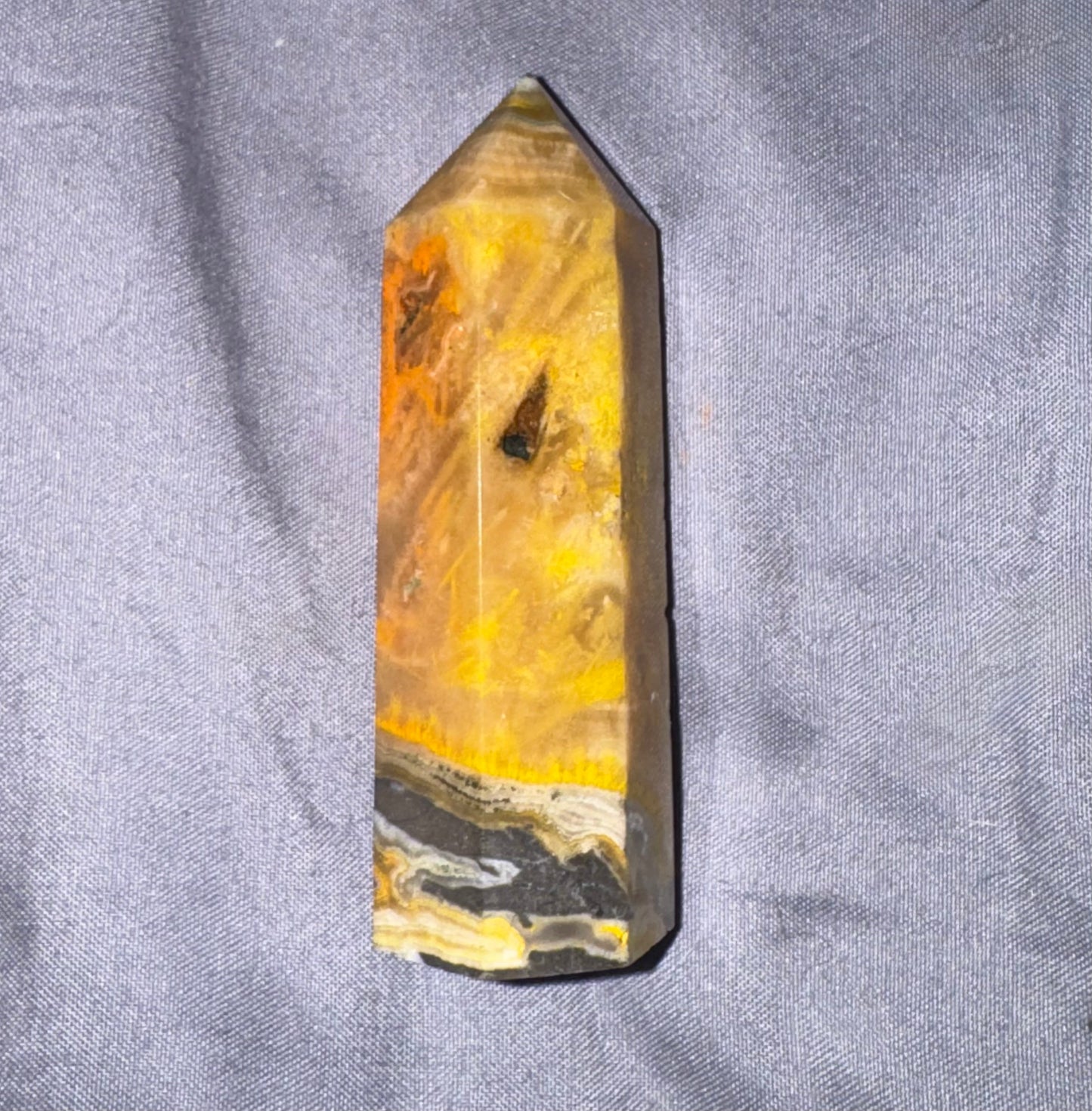 A polished natural quartz crystal point with vibrant yellow, orange, and brown banded patterns and a black inclusions, standing upright on a textured gray fabric background.