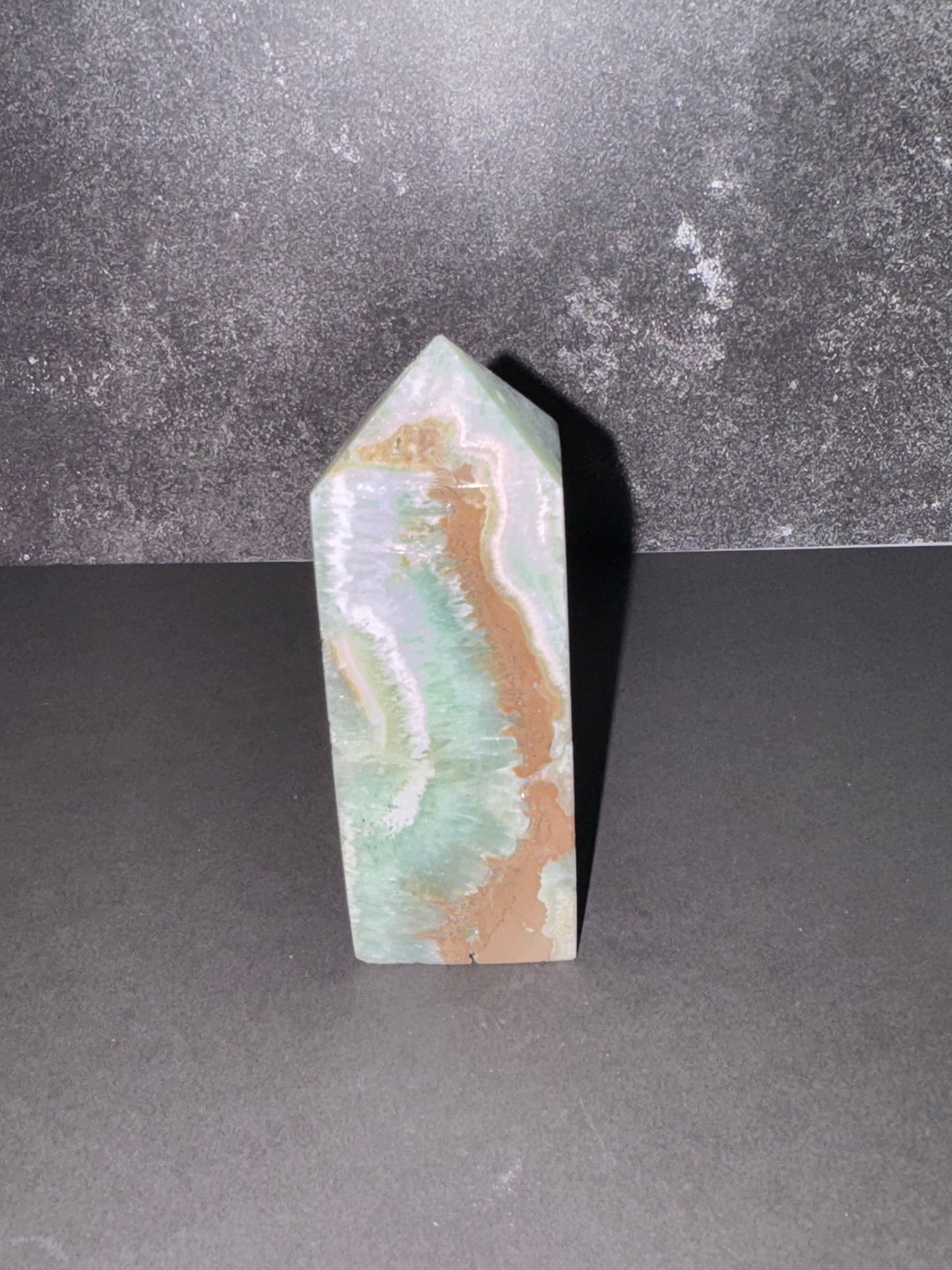 A polished natural crystal point with marbled patterns of light green, beige, and brown, standing upright on a dark gray surface against a textured dark gray background.