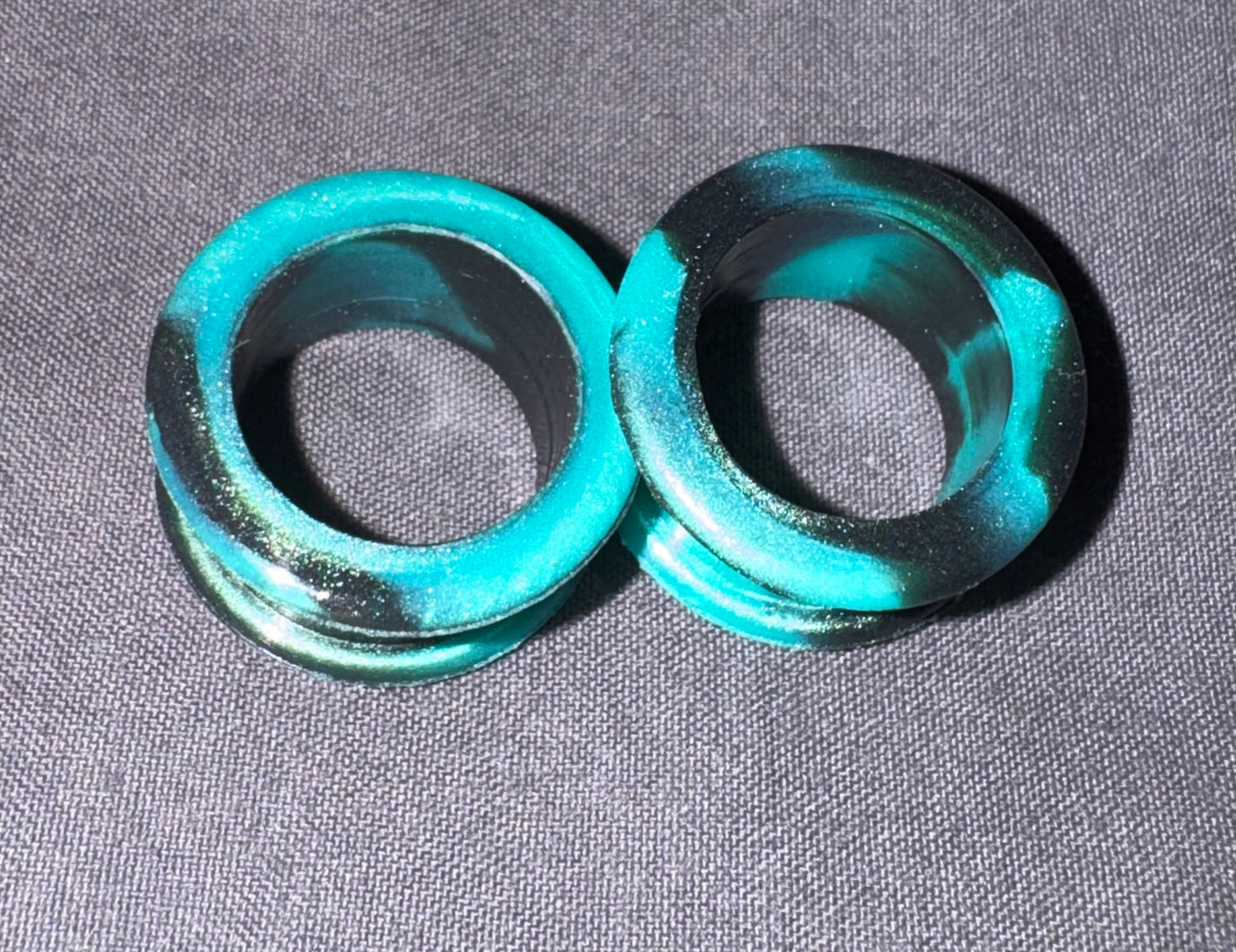 Two circular turquoise and black marbled rings, likely plug-on earrings or decorative rings, displayed on a gray textured fabric background.