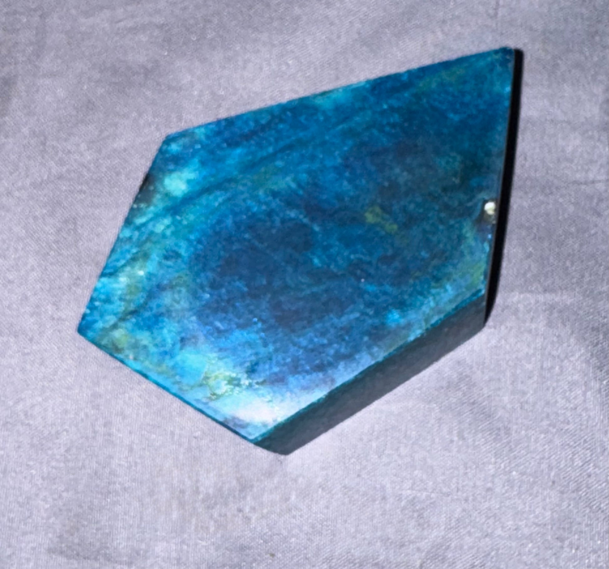 A polished blue hexagonal stone or crystal with natural marbled patterns, resting on a textured gray fabric background.