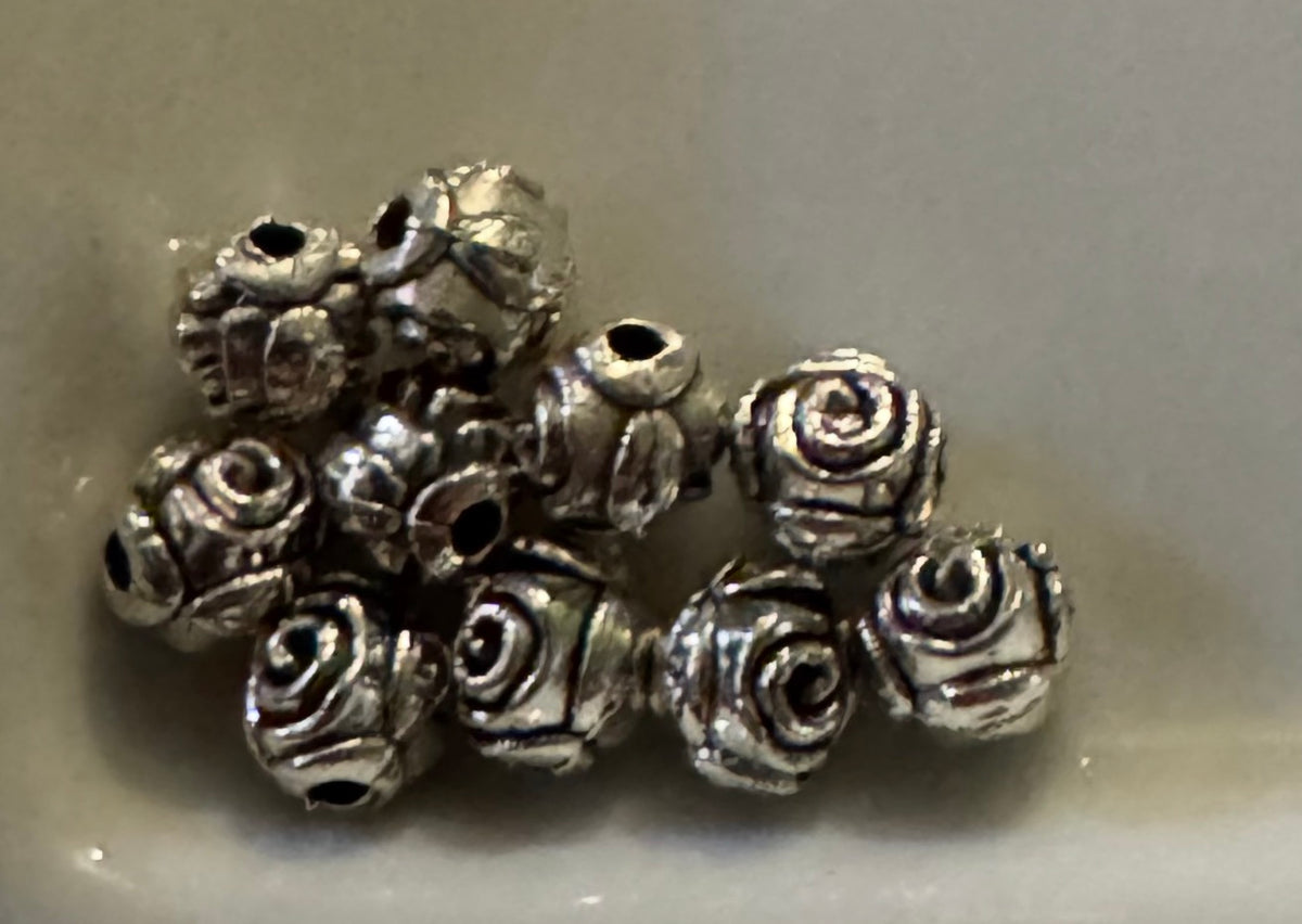 A pile of small metallic rose-shaped beads with a textured, oxidized silver finish, resting on a neutral light gray surface.