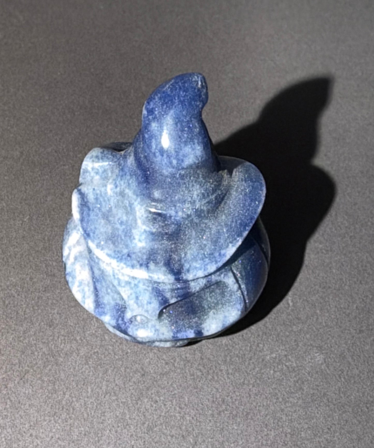 A small blue stone figurine carved to resemble a gnome or elf wearing a pointed hat, photographed on a dark gray surface with a hard shadow cast to the right.
