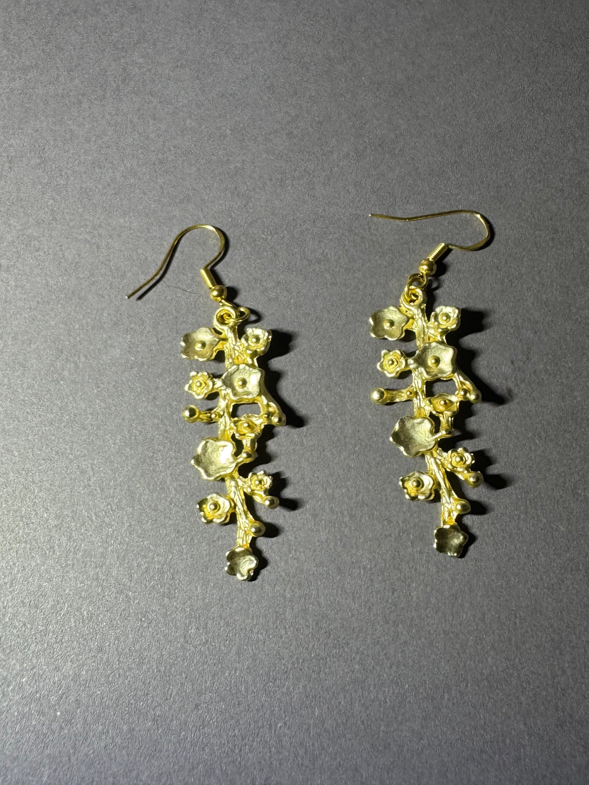 A pair of gold-toned dangle earrings with a floral, branch-like design and small clear rhinestone accents, displayed on a textured gray background.