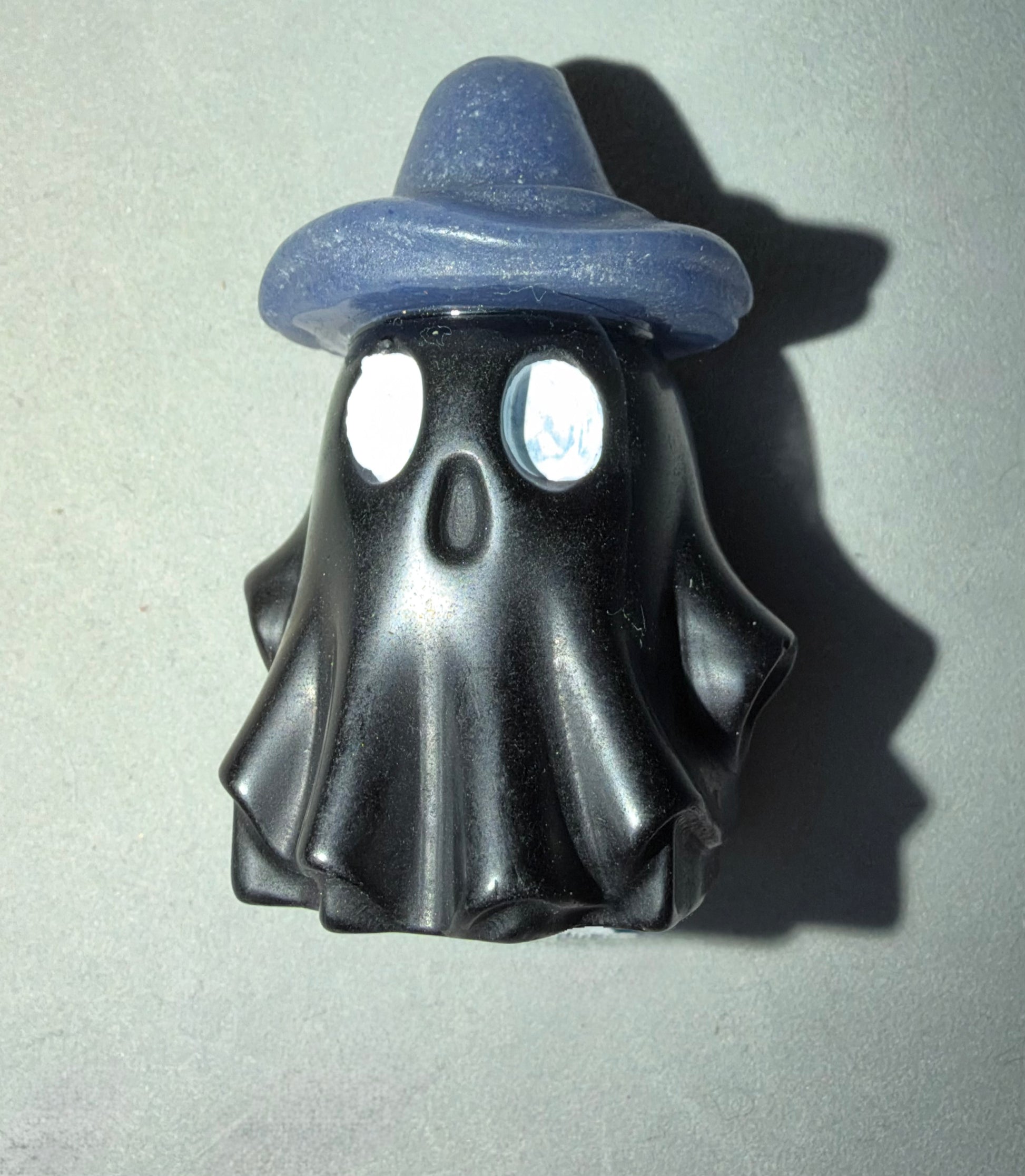 A small ghost-shaped figurine or charm in glossy black with a blue hat, photographed on a light gray background with a strong cast shadow.