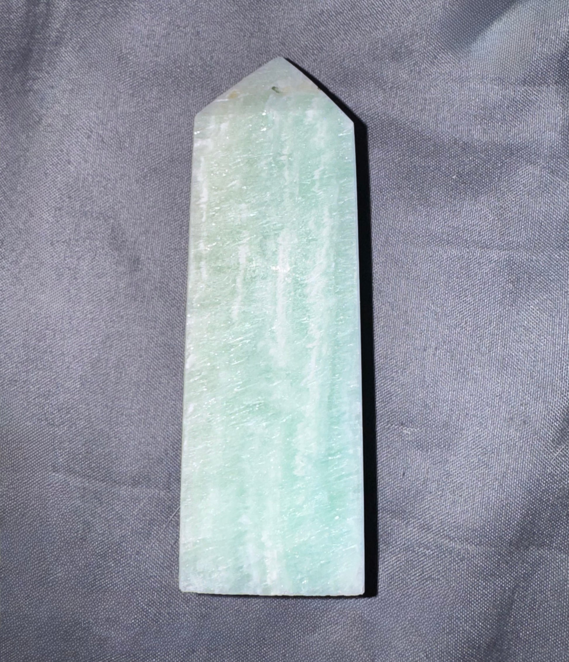 A natural light green amazonite crystal point stake with a smooth, polished surface, standing upright on a textured gray fabric background.