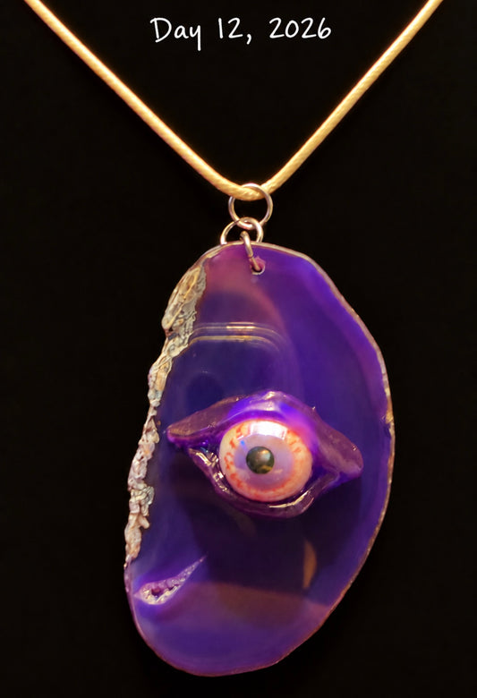 A handmade pendant necklace featuring a purple agate slice with a realistic glass eye charm in the center, suspended from a gold chain against a black background. White handwritten text at the top reads 'Day 12, 2026'.
