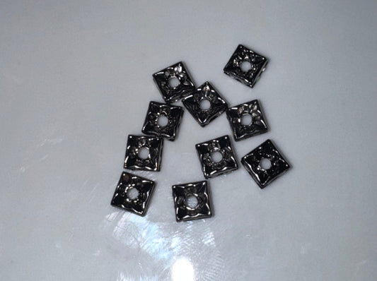 Ten small square metallic charms with a central hole, featuring a textured, hammered finish, arranged on a light gray reflective surface.
