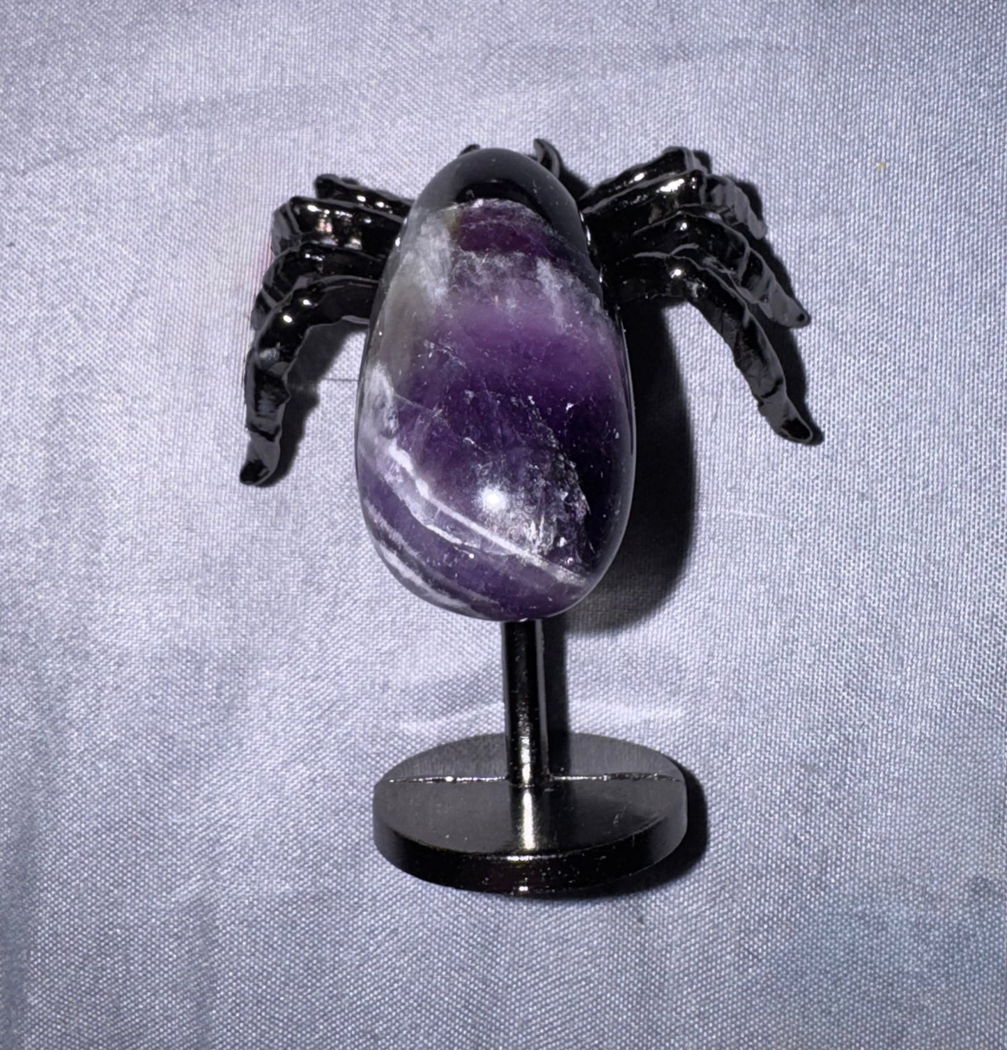 A polished purple amethyst egg-shaped stone displayed on a black spider-shaped stand against a light gray fabric background.