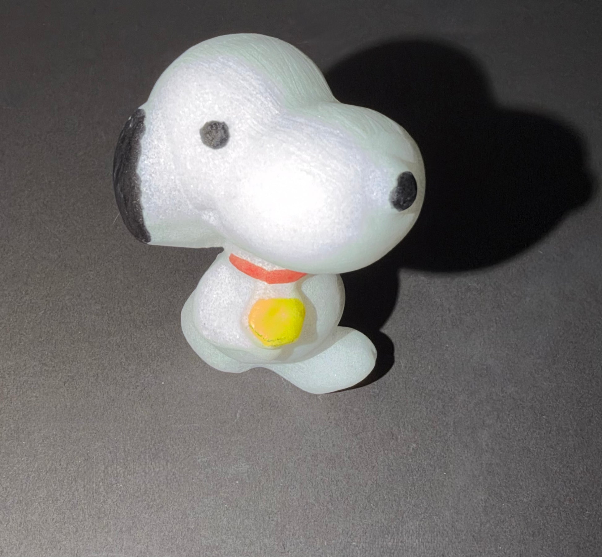 A small white dog figurine with black ears, black eyes, a red collar, and a yellow bell, photographed on a dark gray surface with a strong shadow.