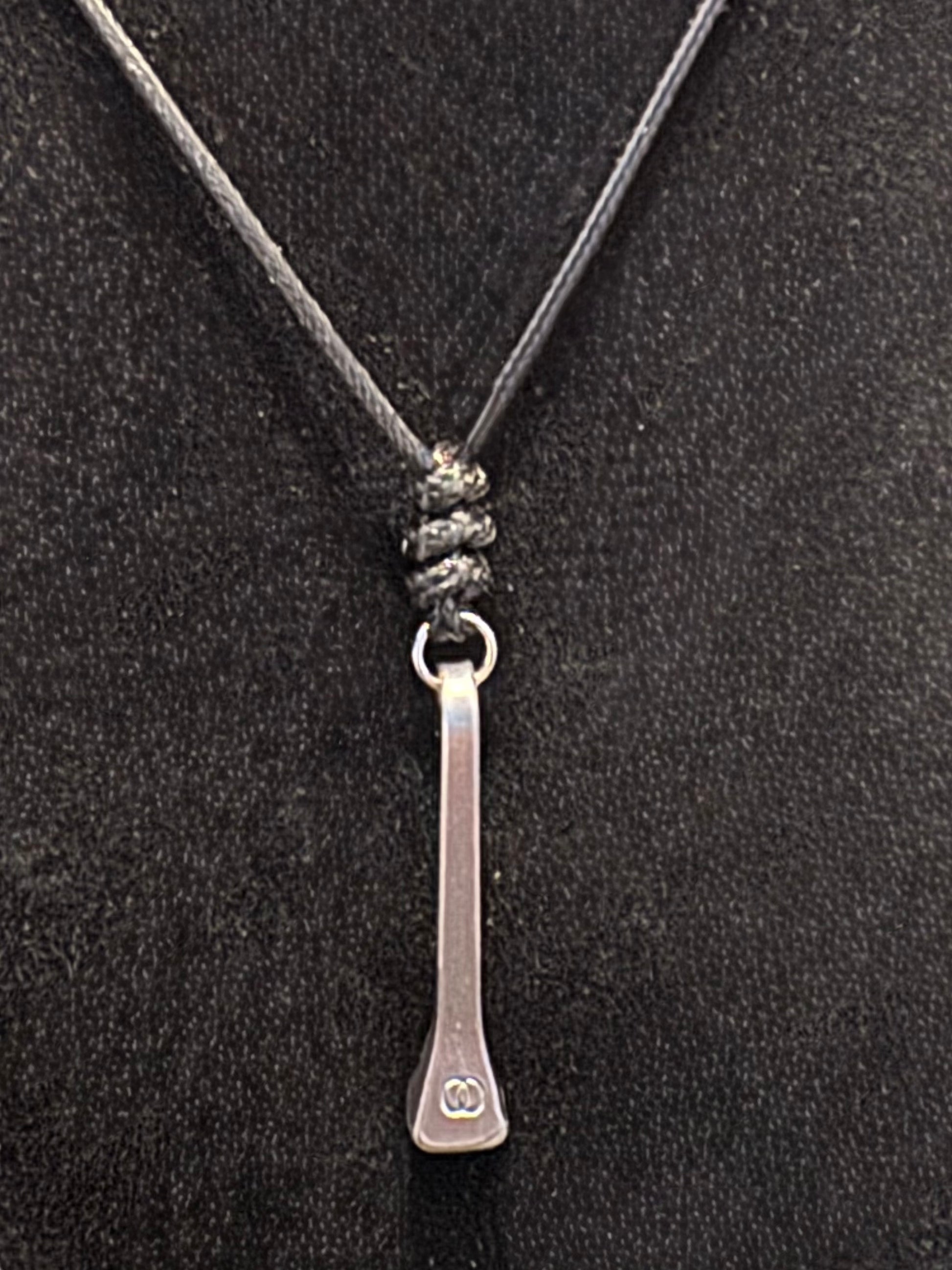 A silver-toned metal pendant necklace with a flat, elongated bar design and a small engraved double-circle logo near the bottom, suspended on a dark gray cord against a black textured background.
