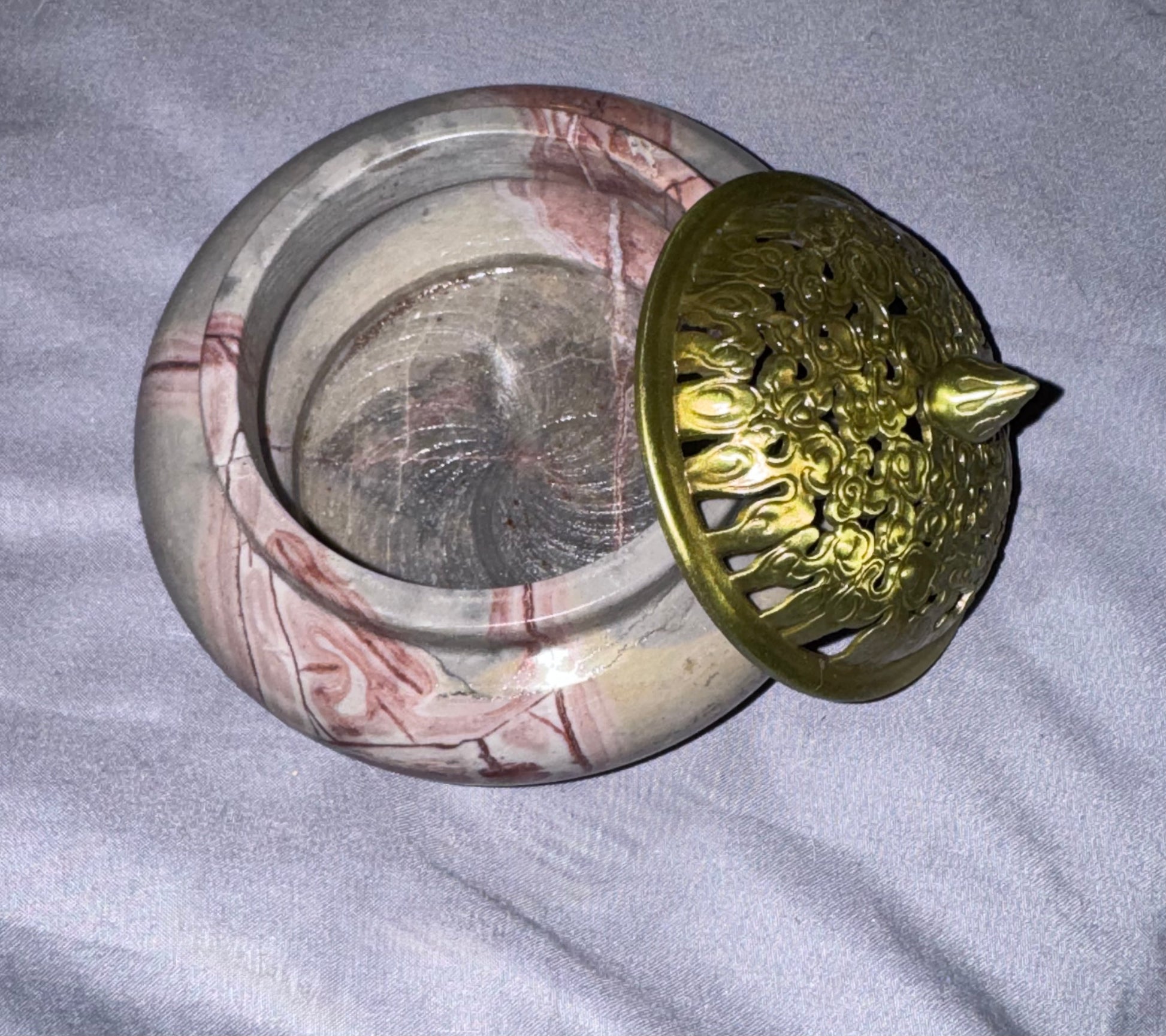 A round stone incense burner with a marbled beige and reddish-brown pattern, shown open with a decorative gold-toned metal lid resting on top, placed on a light gray fabric surface.